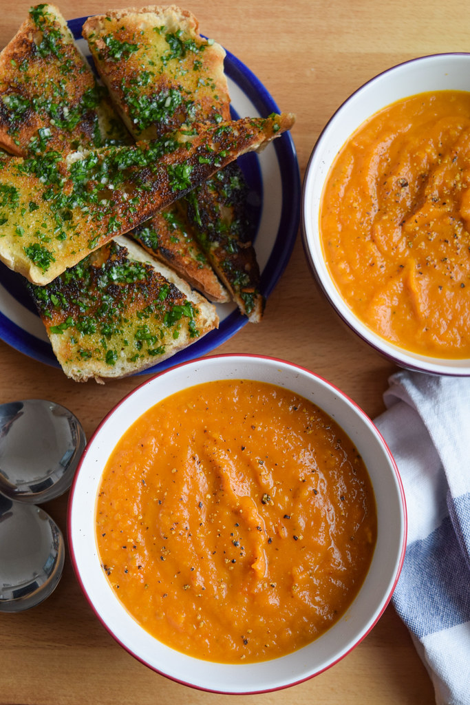 Overhead view of two bowls of tomato and roasted squash soup with a plate of garlic bread to one side.