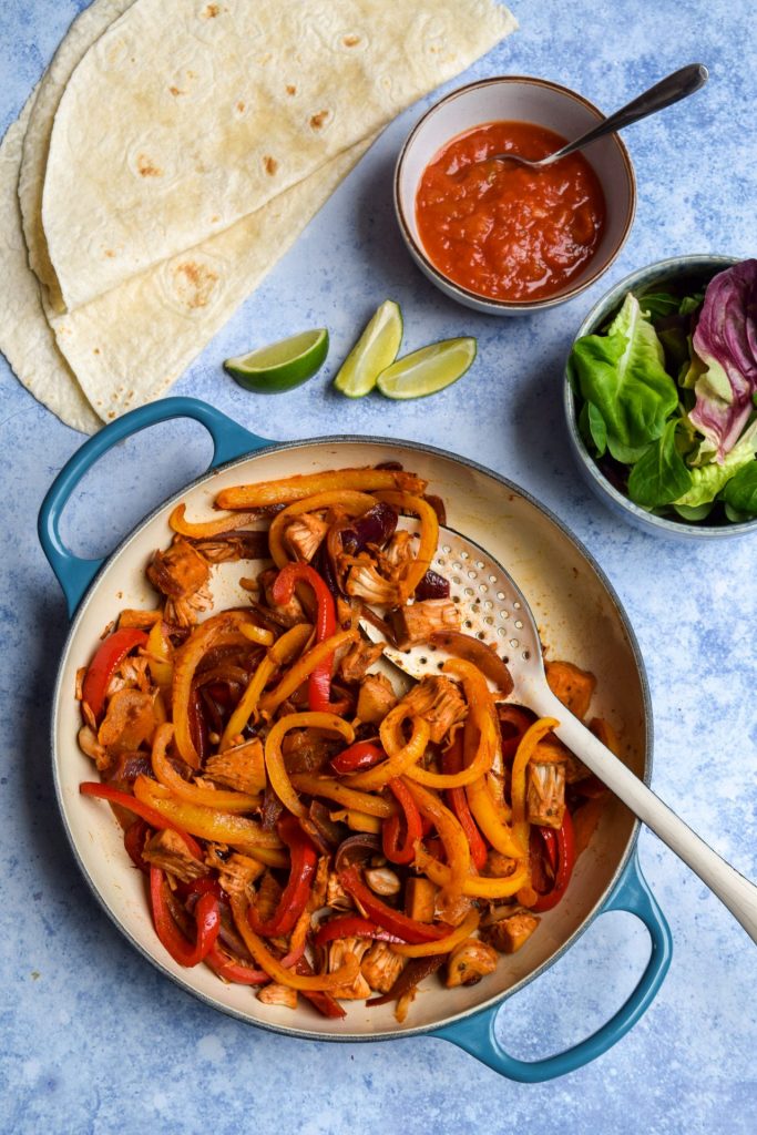 Pan of jackfruit fajitas, wraps and toppings on a blue background.