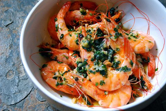 White bowl of prawns in garlic butter with parsley on a grey stone background.