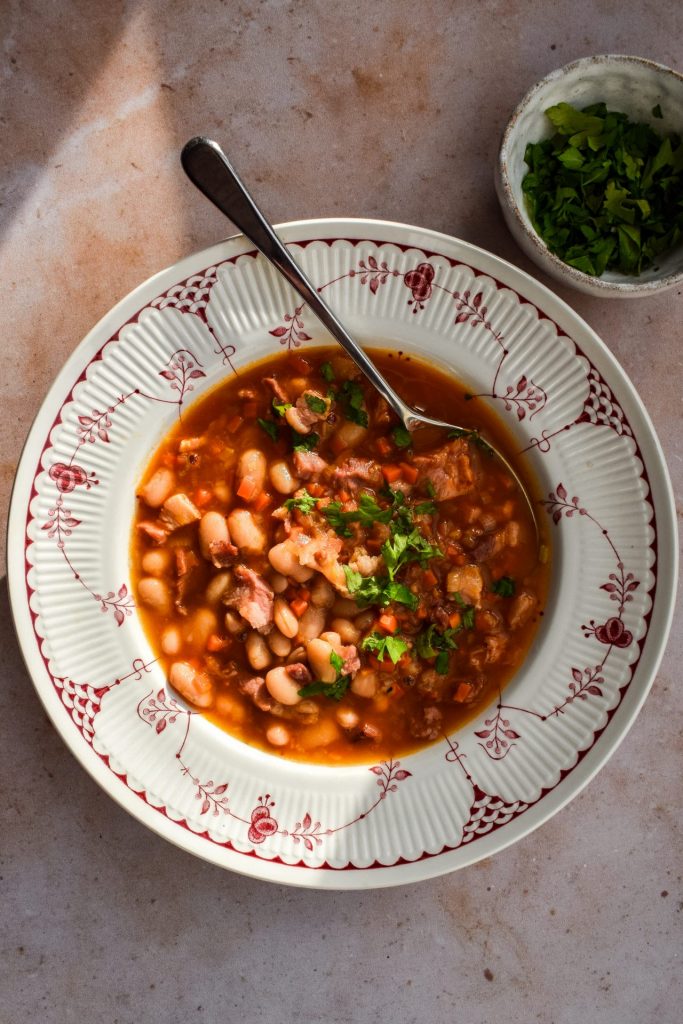 Red and white bowl of bacon and white bean soup with a bowl of flat leaf parsley for scattering off to one side. 