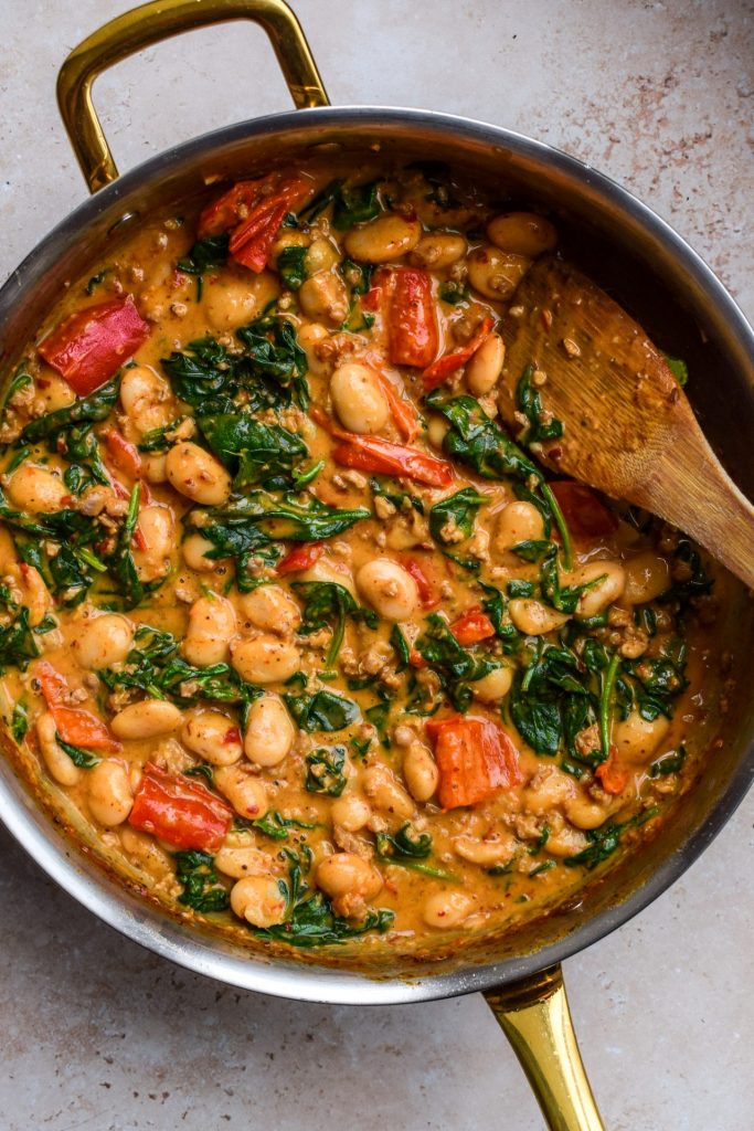 Wooden spatula stirring a skillet of sausage and 'nduja butter beans.