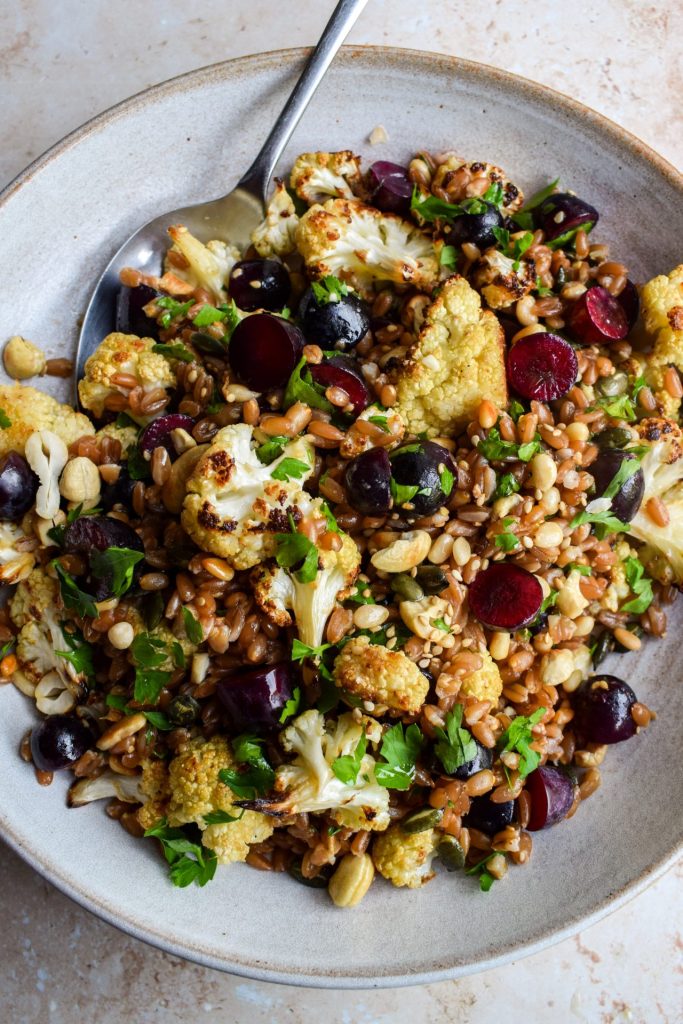 Close up of a grain bowl with roasted cauliflower, parsley, grapes, nuts and seeds mixed in.