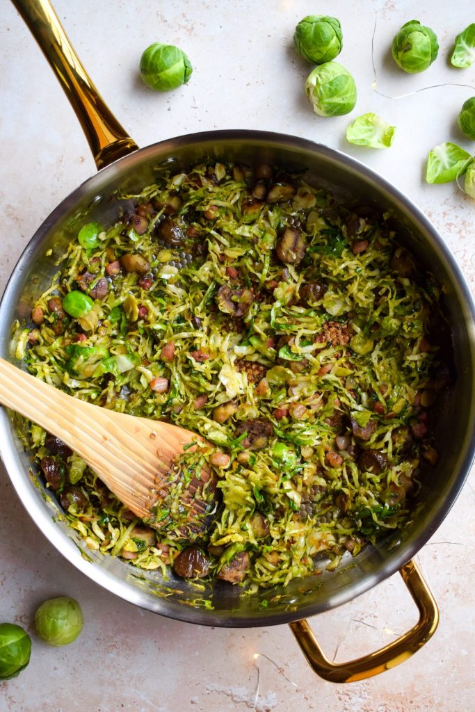 Shredded sprouts with pancetta and chestnuts pictured in a sauté pan with raw sprouts scattered around it.