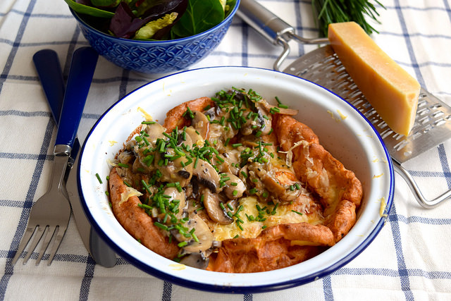 Close up of a german pancake topped with mushrooms and chives with a block of cheese and bowl of salad in the background.