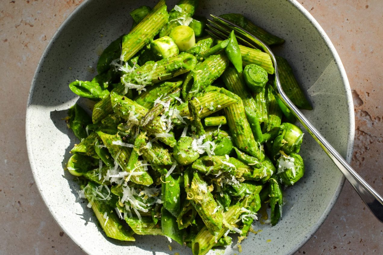 Close up of a bowl of spinach pesto pasta with sliced snap peas, baby leeks and asparagus pictured in direct sunlight.