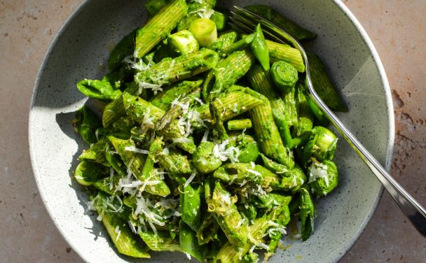 Close up of a bowl of spinach pesto pasta with sliced snap peas, baby leeks and asparagus pictured in direct sunlight.