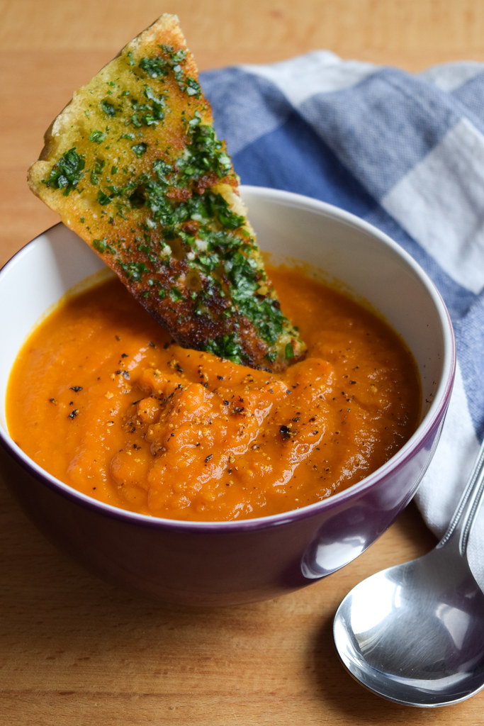 A shard of garlic bread dunked in a bowl of tomato and squash soup.