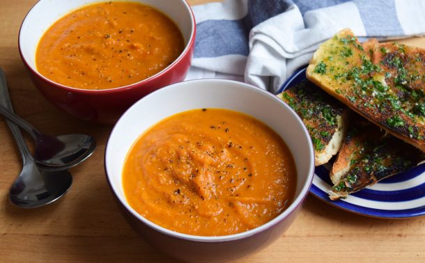 Two bowls of tomato and squash soup with a plate of garlic bread on the side.