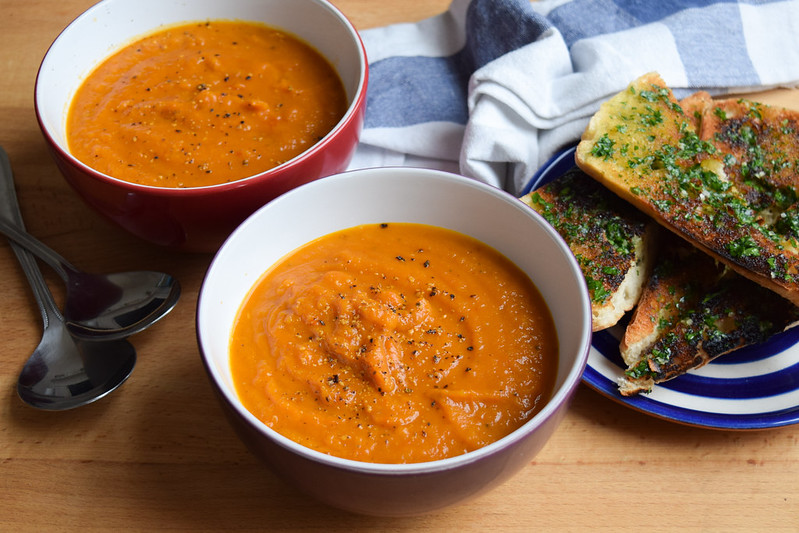 Two bowls of tomato and squash soup with a plate of garlic bread on the side.