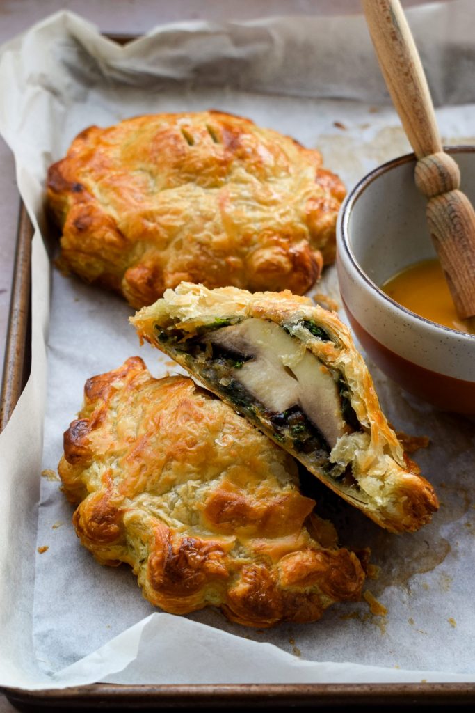 Mushroom wellington sliced in half on a baking sheet with another whole one and a bowl of egg wash in the background.