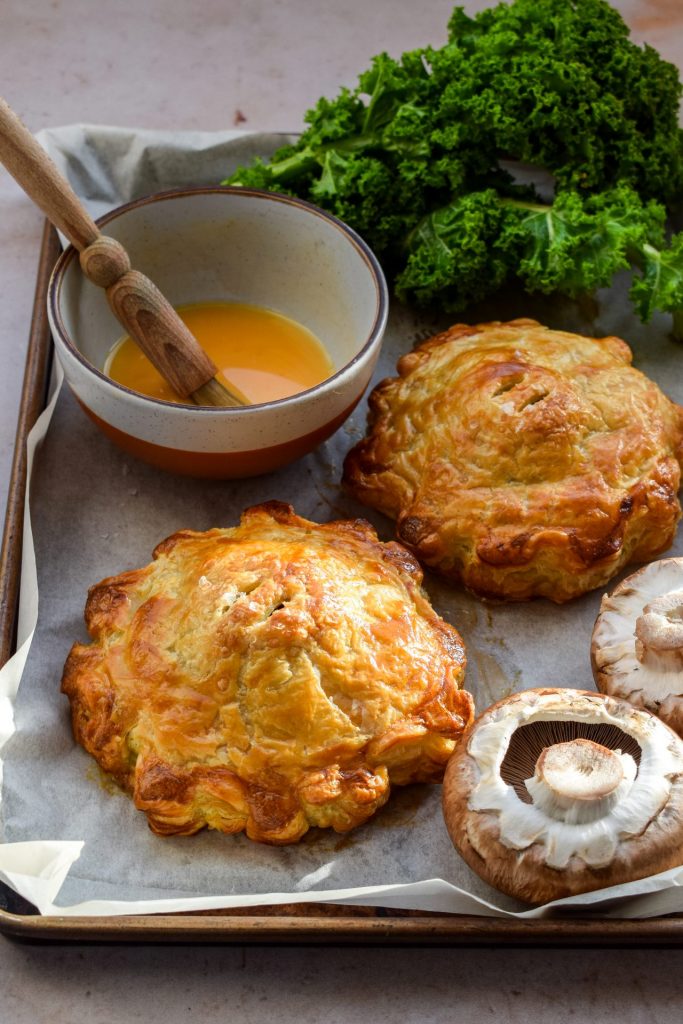 Two portobello mushrooms on a lined baking sheet with a bowl of egg wash, raw mushrooms and kale.