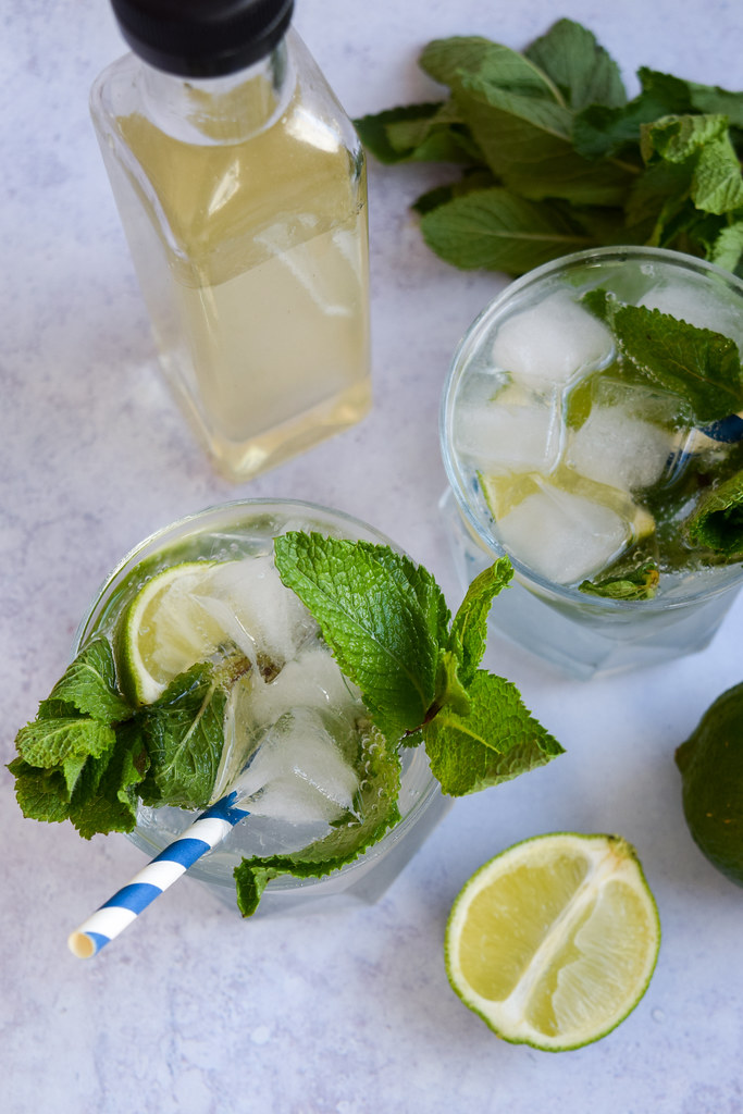 Overhead image of mojitos on a blue background next to a bottle of mojito syrup.