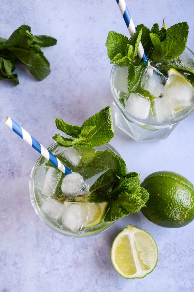 Two mojitos garnished with fresh mint and blue and white striped paper straws on a blue background. There are limes cut and whole next to the glasses.