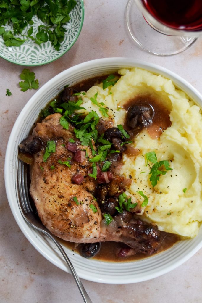 Slow cooker coq au vin with mashed potatoes in a white bowl with a bowl of chopped parsley and a glass of red wine on the side.