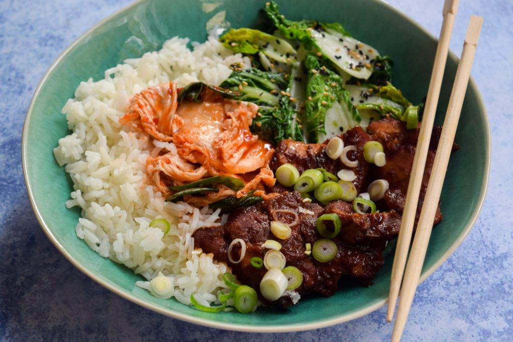 Close up of slow cooker korean chicken thighs with kimchee, sesame pak choi and rice in a blue bowl.