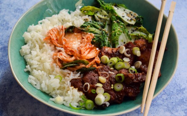 Close up of slow cooker korean chicken thighs with kimchee, sesame pak choi and rice in a blue bowl.