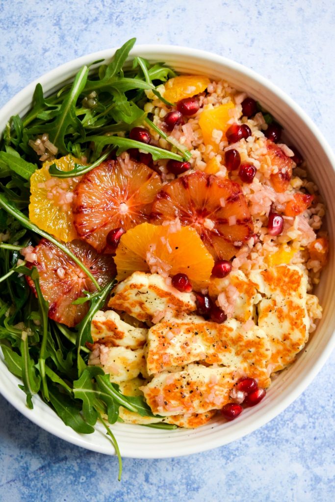 Overhead image of a grain bowl with rocket, sliced oranges, pomegranate and grilled halloumi.