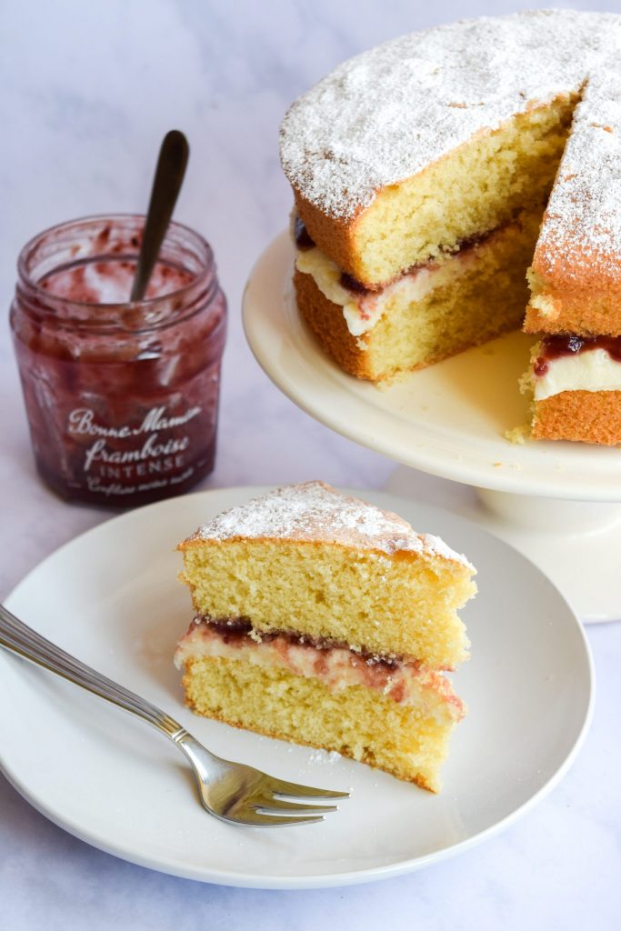 Cake filled with jam and buttercream on a white cake stand with a slice next to it on a white plate, and with a jar of jam in the background.