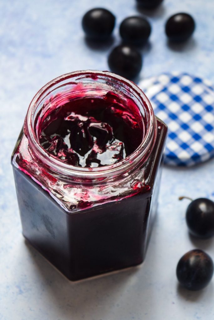 Open jar of jam on a blue background scattered with damsons.