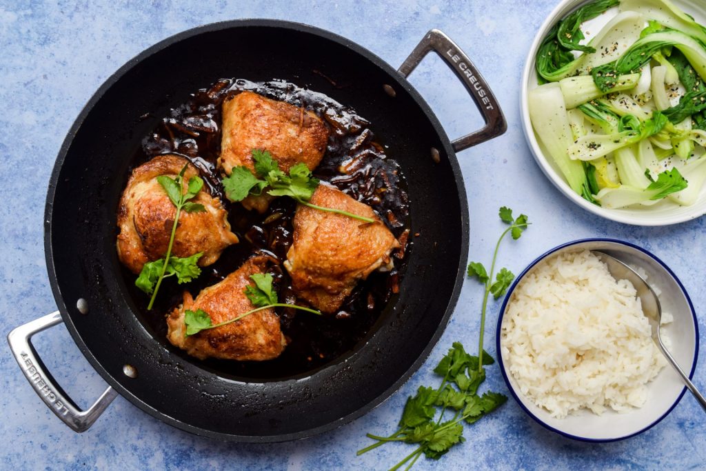 Overhead shot of soy sauce braised chicken thighs in a casserole dish on a blye background with bowls of rice and pak choi on the side.