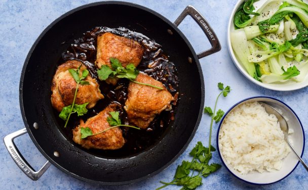 Overhead shot of soy sauce braised chicken thighs in a casserole dish on a blye background with bowls of rice and pak choi on the side.