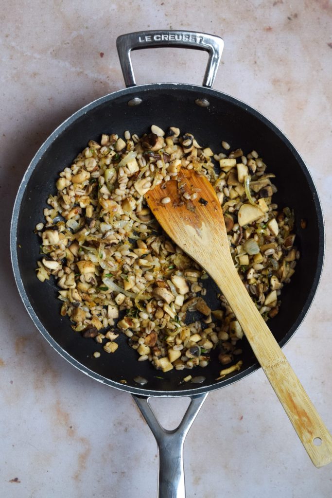 Chopped mushroom and pearl barley filling with a wooden spatula in a Le Creuset Frying Pan.