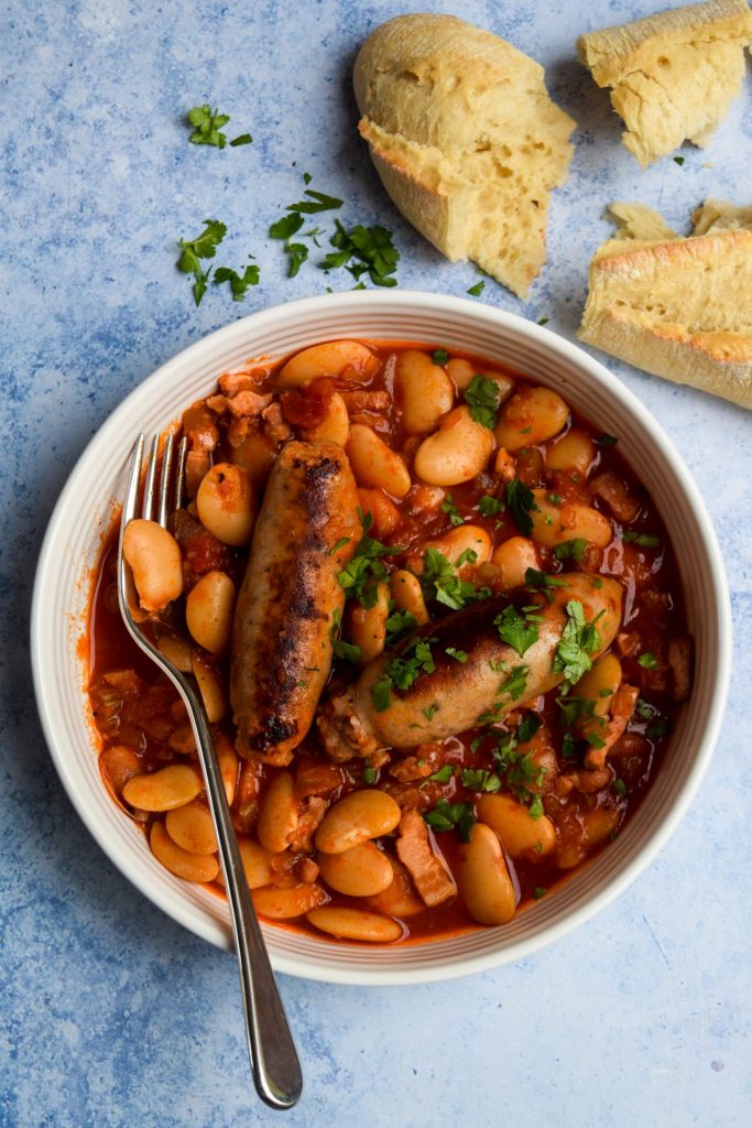 White bowl of sausage and butterbean casserole garnished with parsley on a blue background with a torn baguette. 