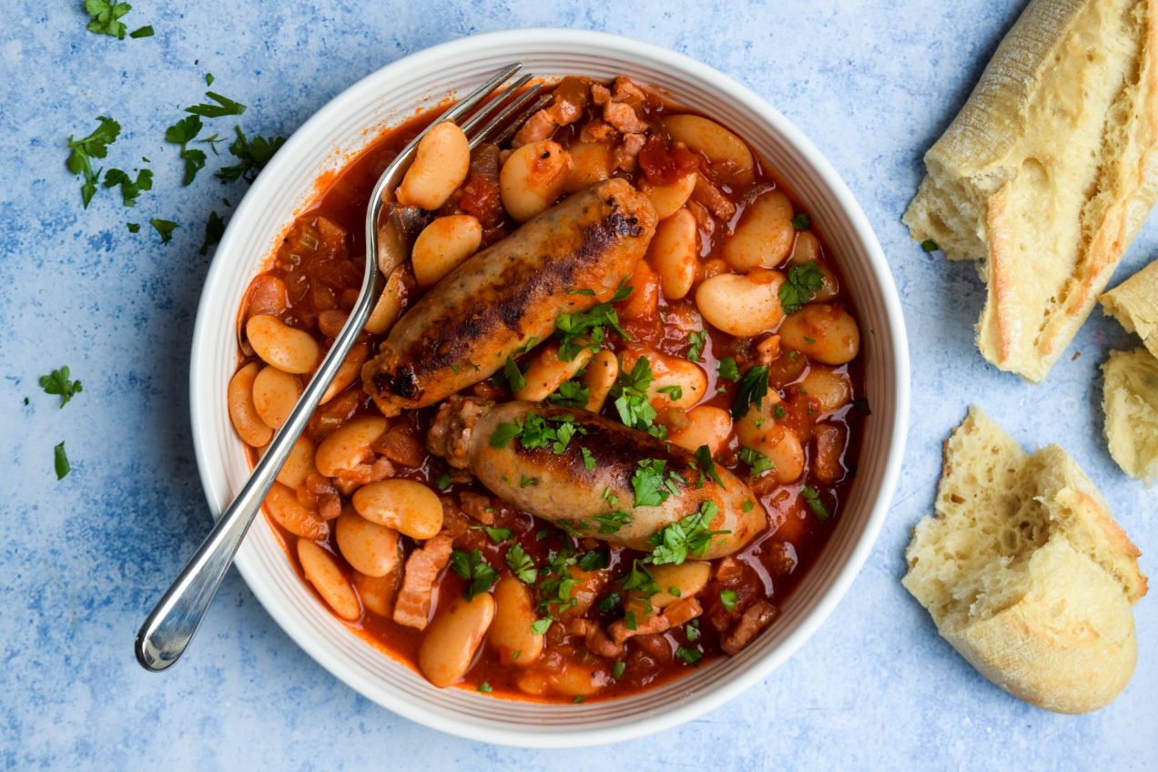 White bowl of slow cooker sausage and butter bean casserole on a blue background.