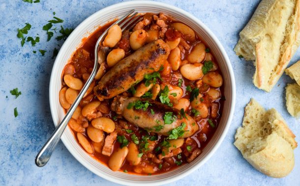 White bowl of slow cooker sausage and butter bean casserole on a blue background.