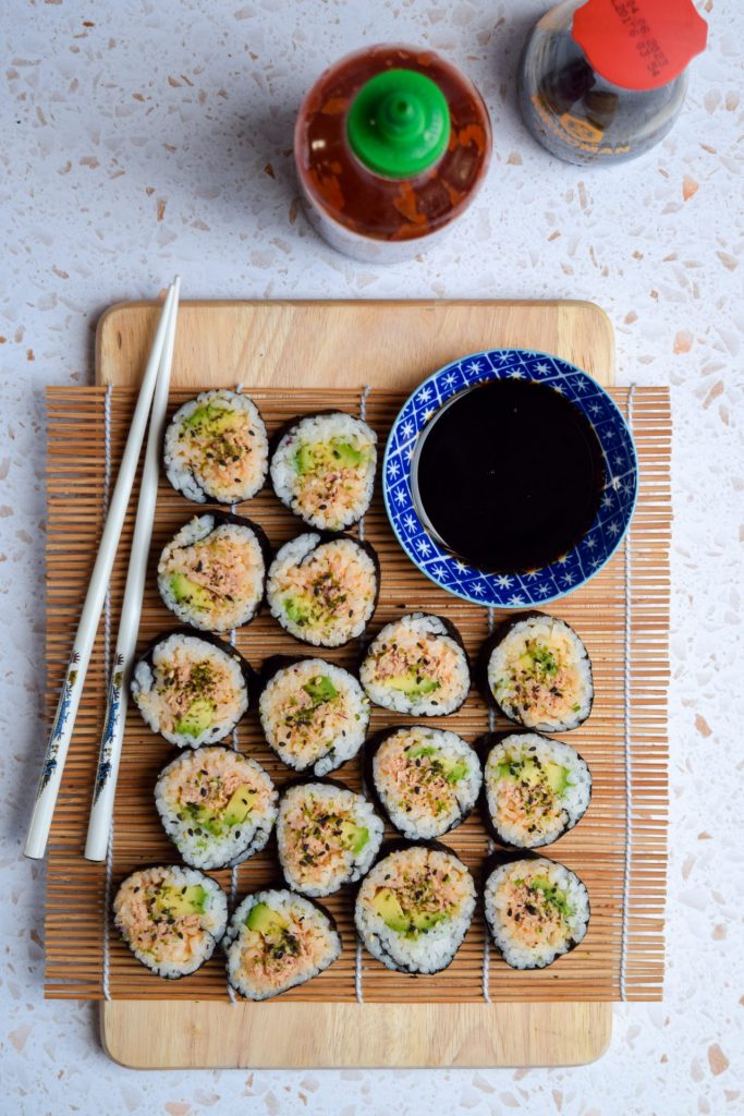 Close up spicy tuna and avocado sushi rolls cut size up on a sushi rolling mat with white chopsticks and a blue dipping bowl of soy sauce.