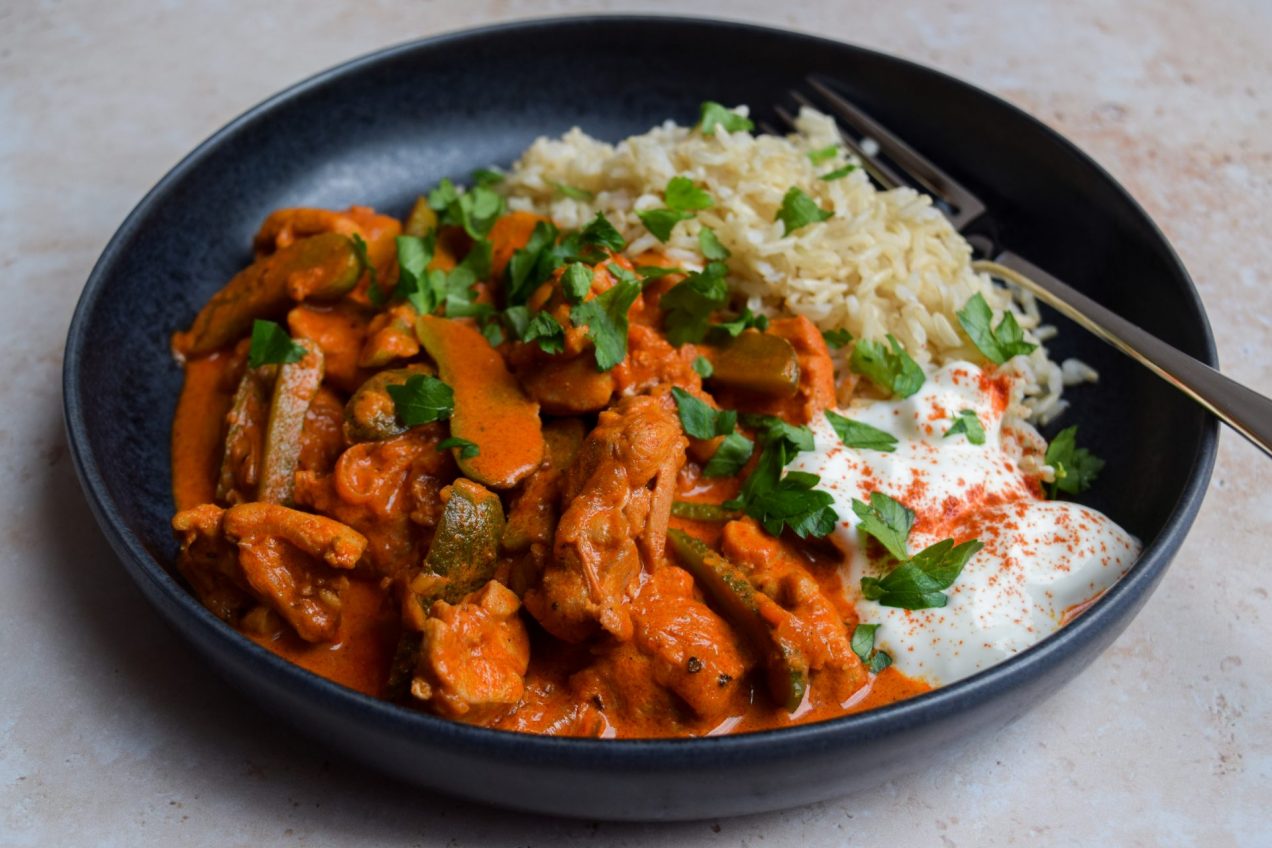 Black bowl of chicken paprikash with sliced gherkins, sour cream and brown rice. Chopped parsley is scattered over the top.