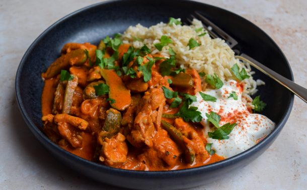 Black bowl of chicken paprikash with sliced gherkins, sour cream and brown rice. Chopped parsley is scattered over the top.