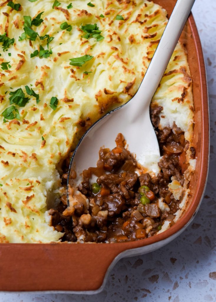 Close up of a spoon lifting a portion of cottage pie out of the baking dish.