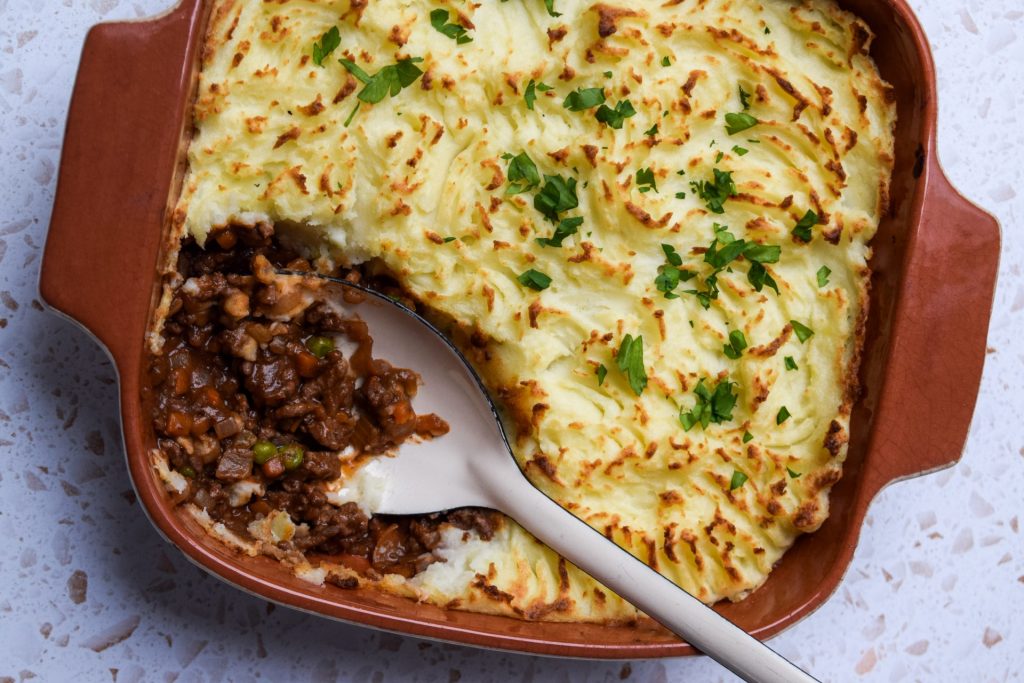 A brown dish of cottage pie with a spoon serving a portion to show the meat filling.