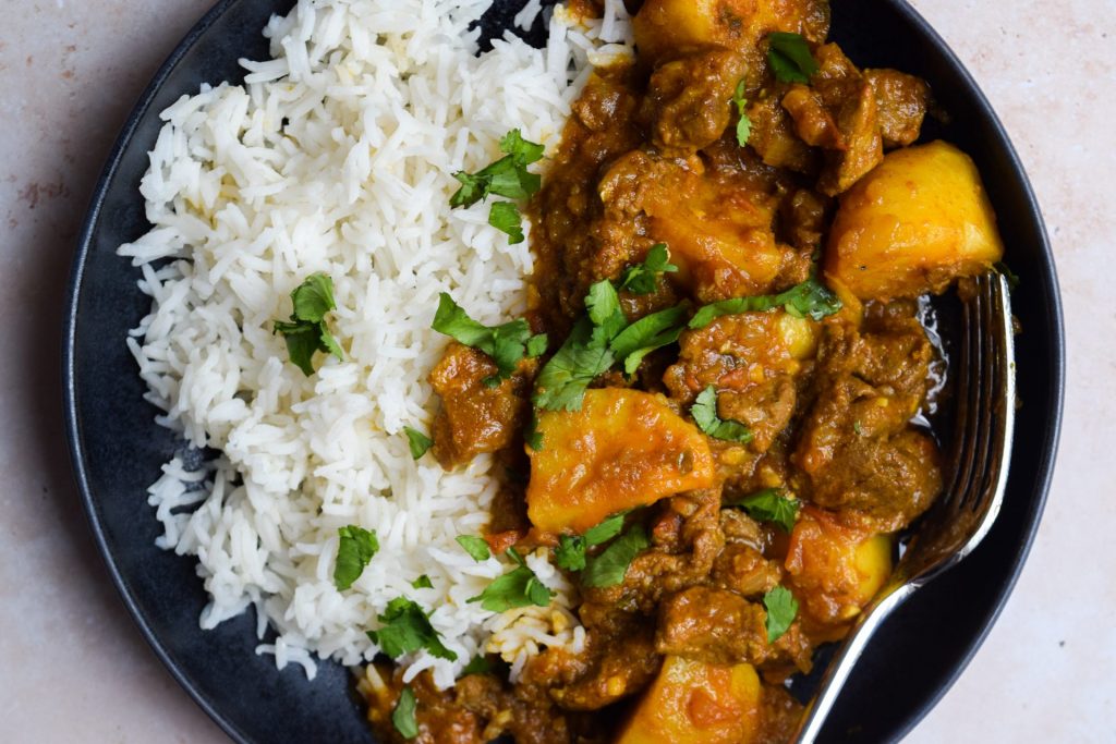 Lamb & Potato Curry (Aloo Gosht) on a black plate with white rice and chopped coriander.