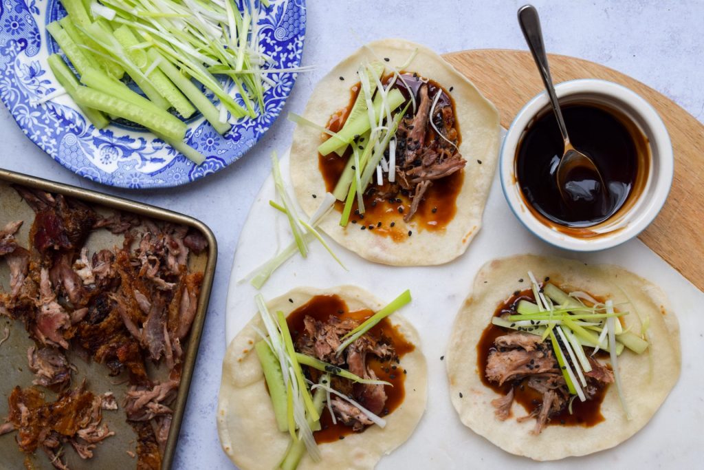 Hoisin Duck Pancake Wraps on a marble board with a bowl of hoisin sauce, blue plate of sliced spring onions and cucumber, and a tray of shredded duck on the side.