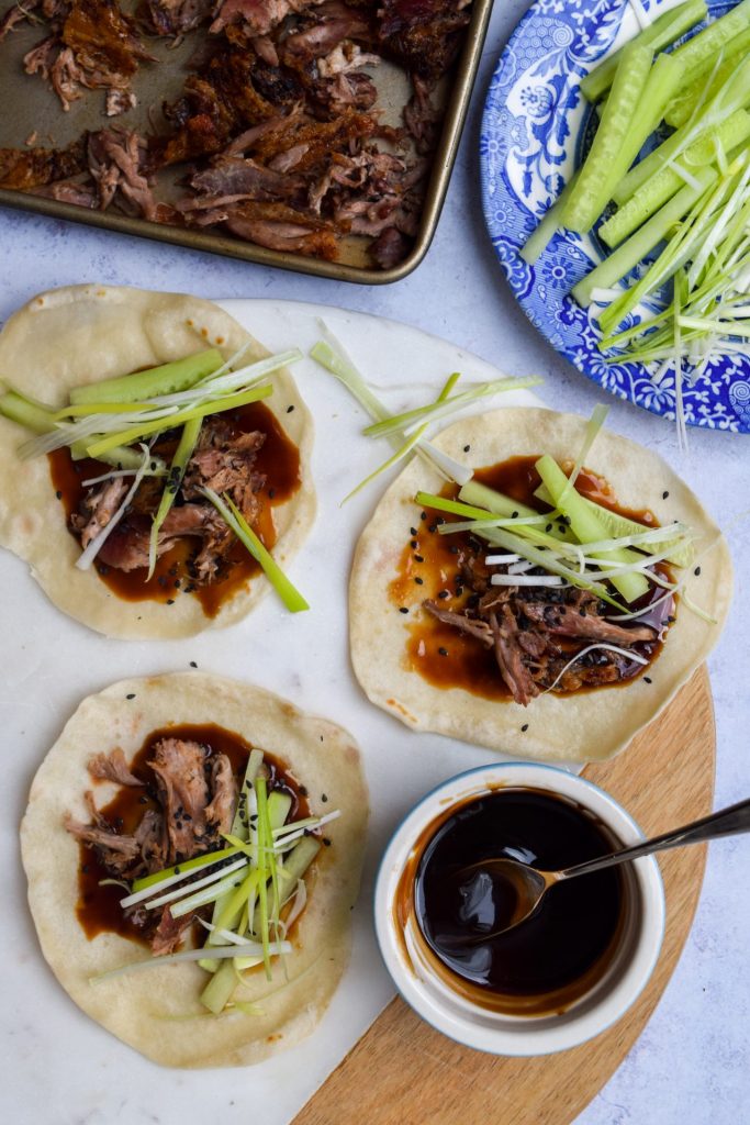 Hoisin Duck Pancake Wraps on a marble board and a light blue background with a bowl of hoisin sauce, blue plate of sliced spring onions and cucumber, and a tray of shredded duck on the side.