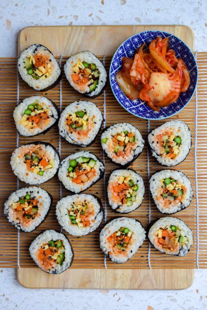 Vegetarian kimbap on a sushi rolling mat with a blue dish of kimchee on the side.