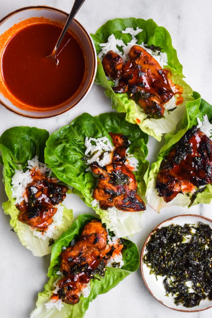 Close up of Korean chicken lettuce wraps with rice, hot sauce and dried seaweed on a marble serving board.