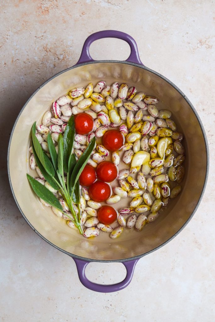 Podded borlotti beans in a casserole dish with cherry tomatoes, sage, water and olive oil.
