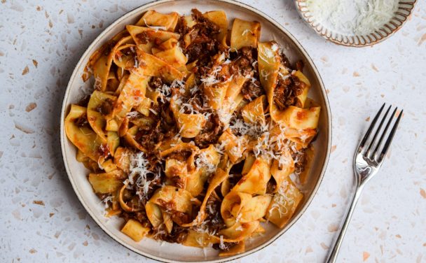 Beef shin ragu tossed with thick noodles in a stone bowl and a bowl of parmesan on the side.
