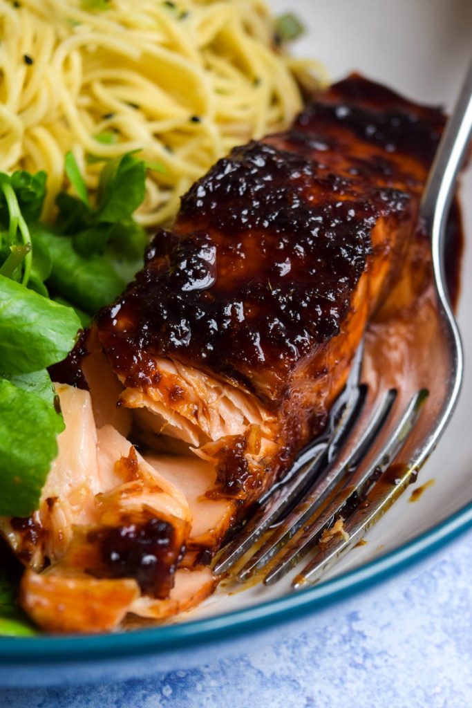 Close up of a piece of oven baked teriyaki salmon being flaked with a fork in a bowl of watercress and sesame noodles.