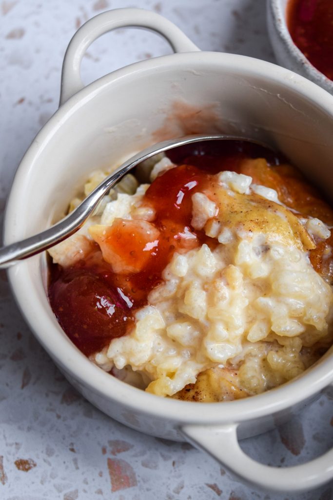 Bowl of baked rice pudding with a silver spoon and a generous dollop of homemade strawberry jam.