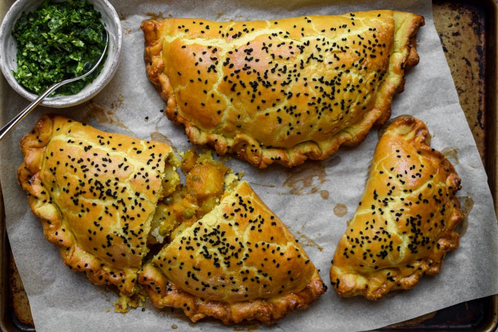 Three curried potato pasties sprinkled with nigella seeds on a baking tray.