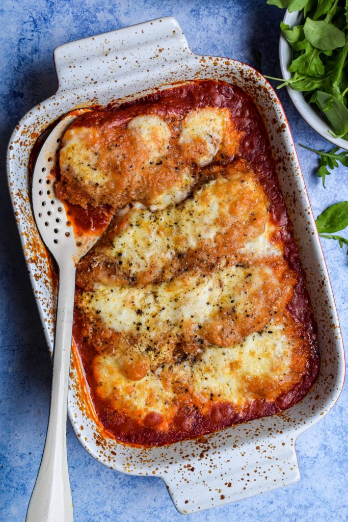 Dish of chicken parmigiana on a blue background with a bowl of rocket on the side. 