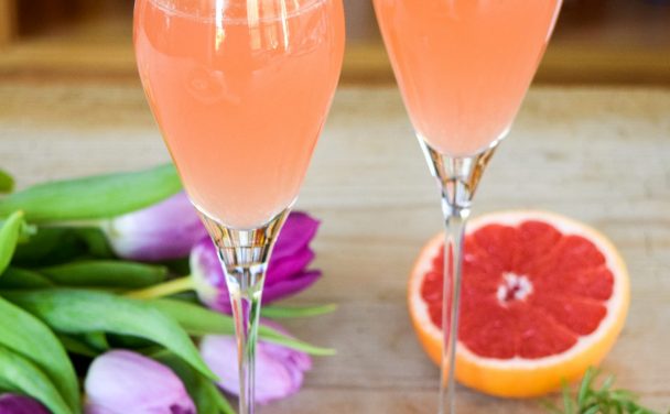 Pair of mimosas on a table with a bunch of tulips and a cut grapefruit in the background.