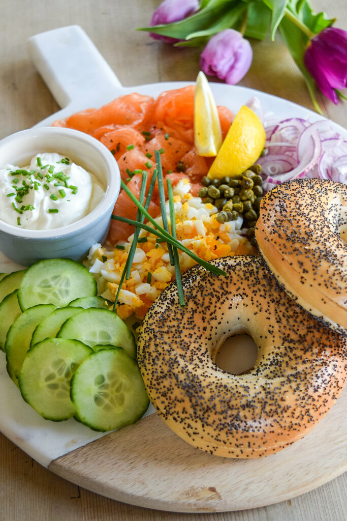 Close up of a bagel board with cucumber, chopped egg, chives, whipped feta, capers, red onion and lemon wedges.