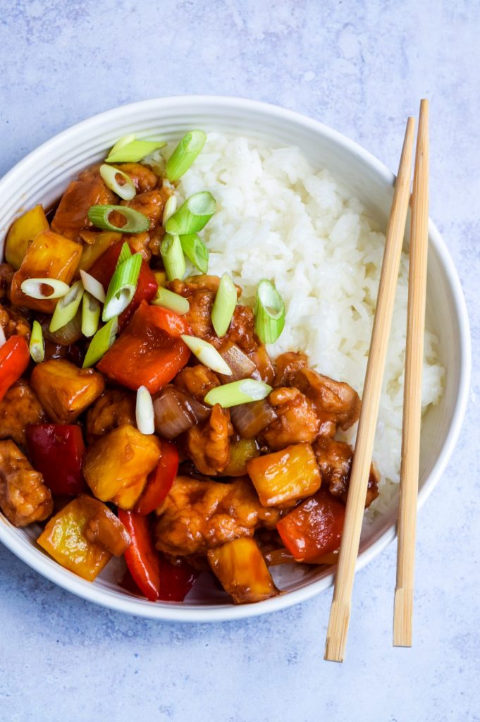 Close up of a serving of sweet and sour chicken and white rice in a white bowl on a blue background.