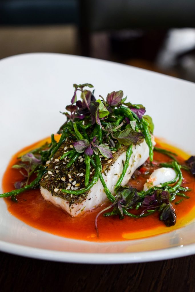 Za'atar crusted cod in a pool of tomato sauce topped with herbs and samphire on a white plate.