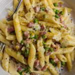 White bowl of penne with peas, pancetta, lemon zest and shaved parmesan.